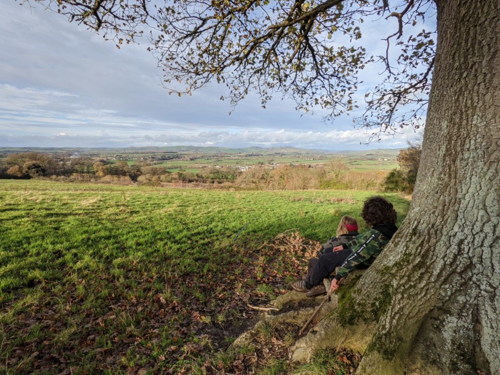 two-boys-sat-under-a-tree-looking-out-across-green-fields-in-the-distance.