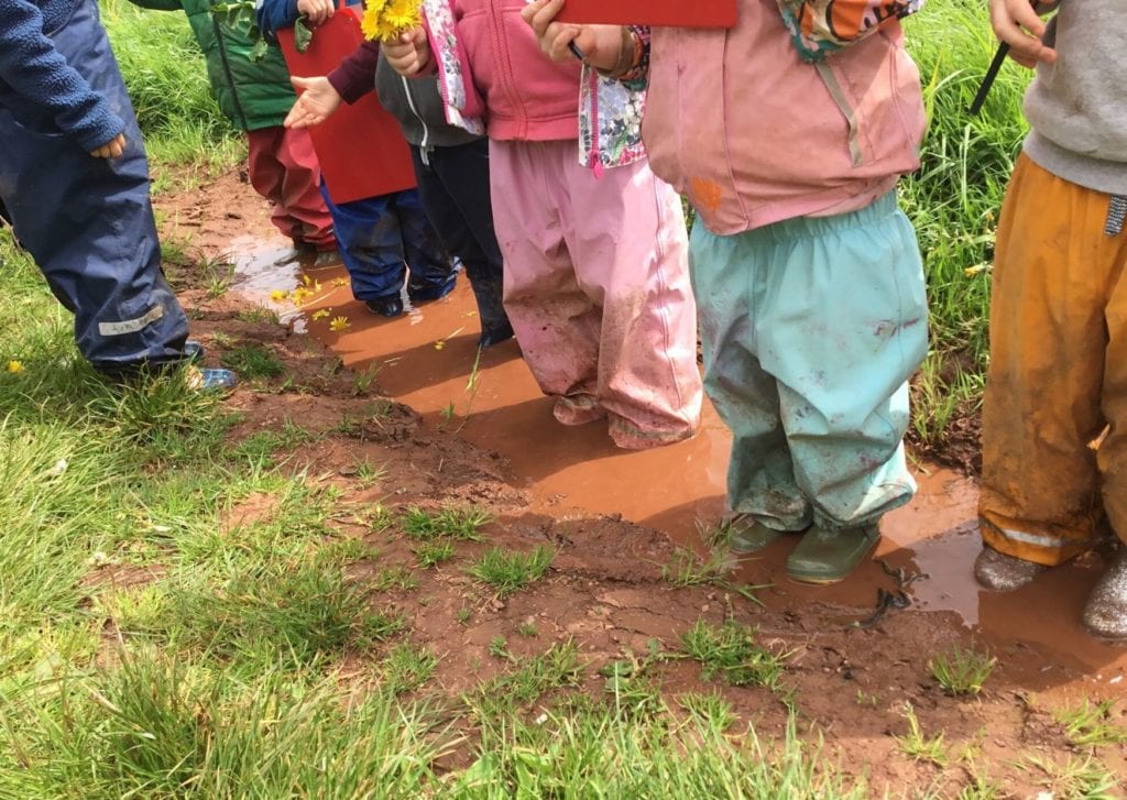 A-row-of-children-wearing-waterproofs-and-welly-boots-stand-in-a-muddy-puddle-in-a-field.